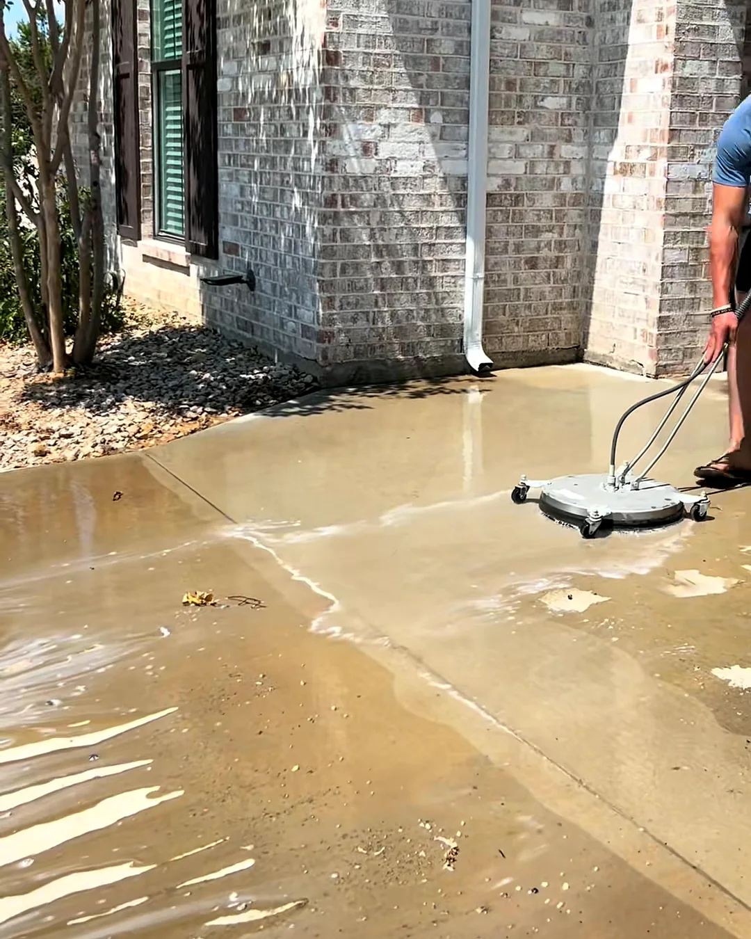 Landon running a surface cleaner across a dirty concrete patio beside a brick house